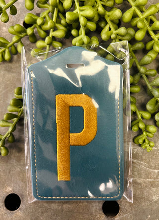 Embroidered monogram luggage tag in clear protective packaging surrounded by green foliage decoration.