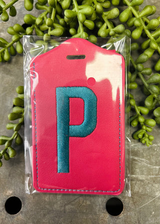 Red leather luggage tag with embroidered letter P in clear plastic packaging on natural background