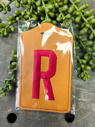 Embroidered monogram luggage tag in clear plastic packaging surrounded by green foliage on stone surface.