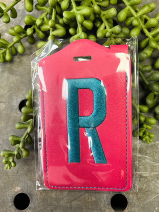 Red leather luggage tag with embroidered letter R in clear plastic packaging surrounded by greenery