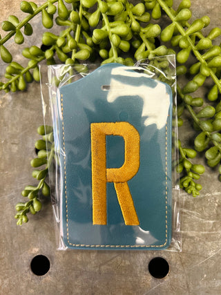 Embroidered monogram luggage tag in clear packaging surrounded by decorative greenery on marble surface