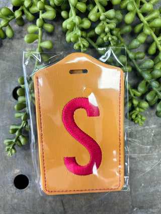 Leather luggage tag with embroidered monogram letter in clear protective packaging surrounded by green foliage