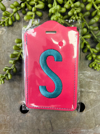 Monogram luggage tag with embroidered letter S in clear plastic packaging on stone surface with greenery.