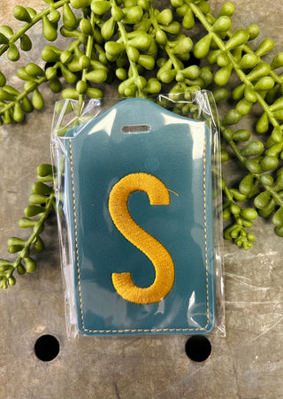 Monogram luggage tag with embroidered letter in clear protective packaging surrounded by green foliage