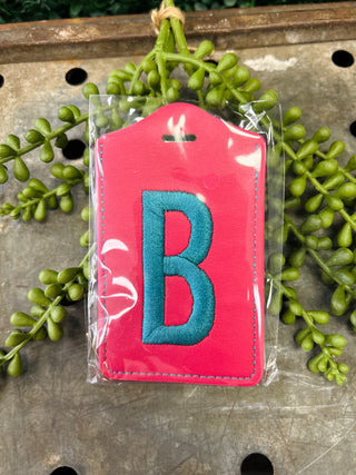 Pink monogram luggage tag with embroidered letter B in clear protective packaging on wooden surface