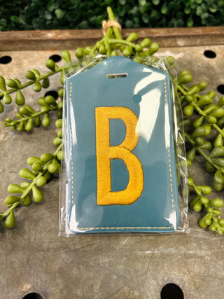 Blue leather luggage tag with embroidered letter B in yellow, packaged in clear plastic wrap.
