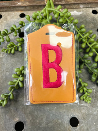 Monogram luggage tag with embroidered letter in clear packaging surrounded by green foliage
