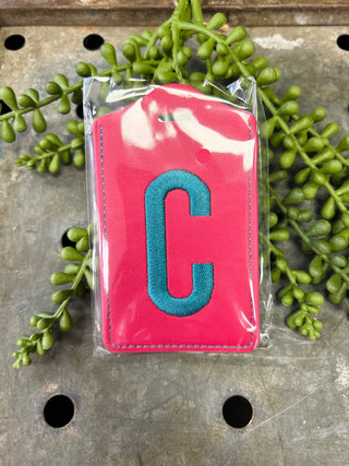 Pink leather luggage tag with embroidered letter C in clear protective packaging surrounded by green plants.