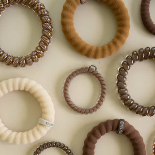 Collection of TELETIES spiral and fabric hair ties in warm neutral tones arranged on a light surface.