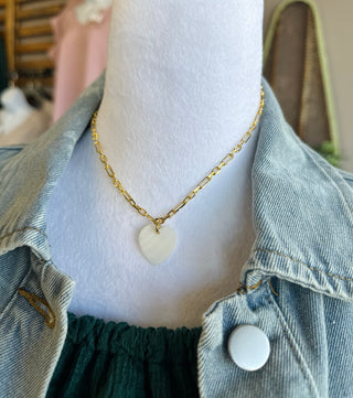 Gold chain necklace with white heart pendant displayed on mannequin wearing denim jacket and green top.