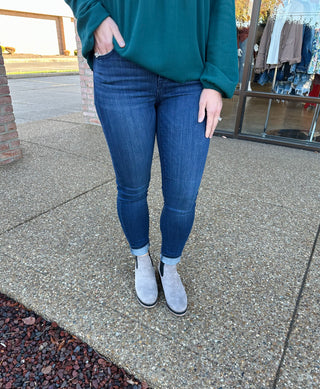 Person wearing dark wash skinny jeans with teal sweater and gray ankle boots standing on concrete sidewalk.