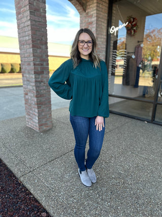 Woman wearing dark wash skinny jeans with teal blouse and ankle boots in outdoor shopping area.