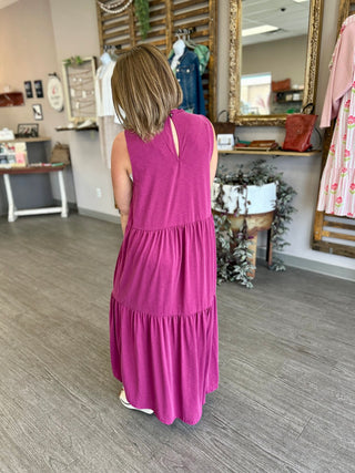 Woman wearing a sleeveless tiered maxi dress in bright pink, shown from behind in boutique setting.