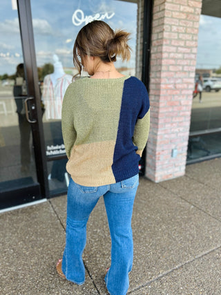 Woman wearing a colorblock knit sweater with flare jeans, shown from behind outside a store.