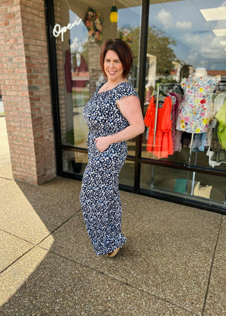Woman wearing navy floral print jumpsuit standing outside clothing boutique storefront with displayed garments.