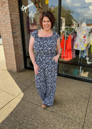 Woman wearing navy floral print jumpsuit with ruffle sleeves standing outside clothing boutique storefront.