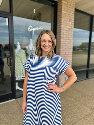 Woman smiling in a striped t-shirt dress with chest pocket, posing outside boutique.