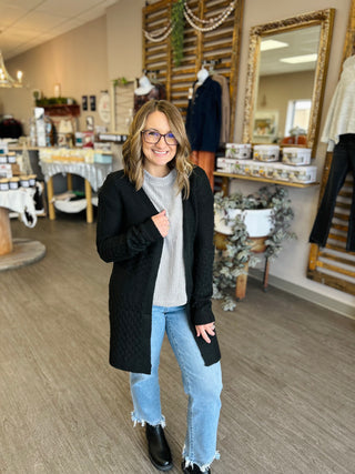 Woman wearing black knit cardigan with cable detail over gray sweater and jeans in boutique setting.