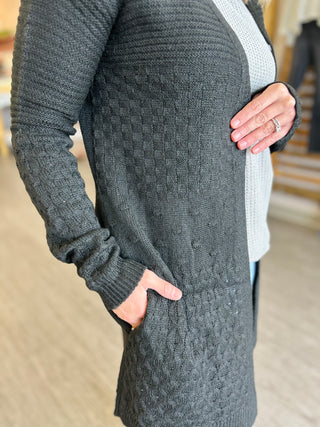 Woman wearing gray cable-knit cardigan with pockets and detailed texture, hands in pockets.