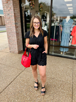 Woman wearing black short-sleeve tie-waist romper with scalloped hem, standing outside boutique storefront.