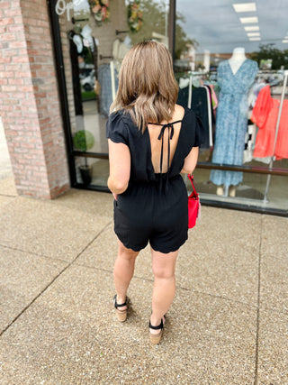 Woman wearing black short romper with open tie-back detail and platform sandals outside boutique storefront.