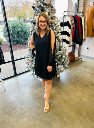 Woman wearing black sleeveless dress with sheer overlay in clothing boutique with Christmas tree backdrop.
