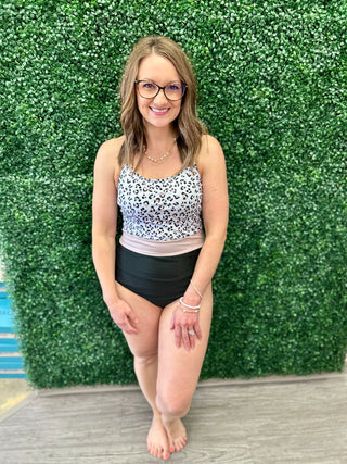 Woman wearing leopard print one-piece swimsuit with black bottom, posing against green hedge backdrop.
