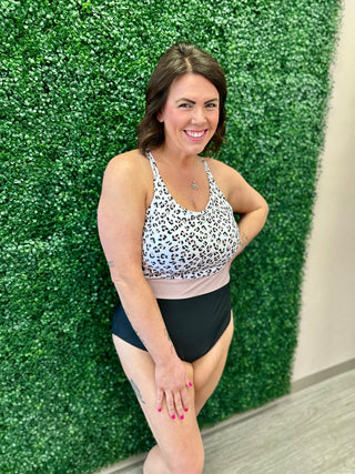 Woman wearing leopard print one-piece swimsuit with color-blocked design, posing against green hedge backdrop.