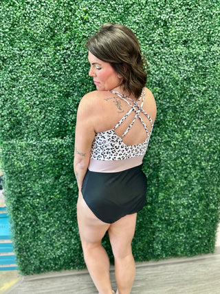 Woman wearing leopard print halter one-piece swimsuit with criss-cross back straps against green hedge backdrop.