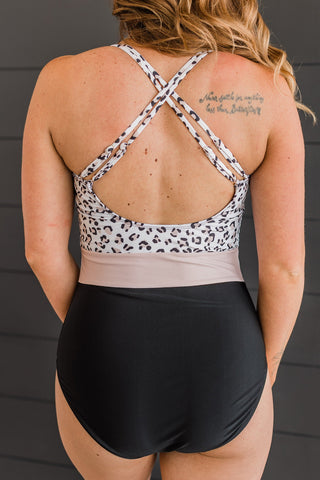 Woman wearing leopard print and black one-piece swimsuit with cross-back straps, shown from behind.