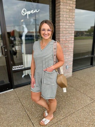 Woman wearing a striped sleeveless romper with front pockets, paired with white sandals and a tan bag.