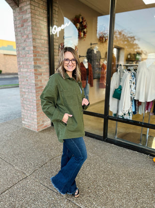 Woman wearing olive green button-up jacket with pockets and blue jeans standing outside clothing boutique.