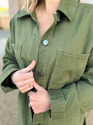 Woman wearing olive green denim jacket with chest pockets and button closure, showing relaxed fit styling.