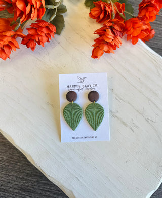 Handcrafted polymer clay leaf dangle earrings with dark round studs displayed on a brand card.