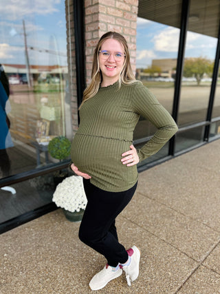 Pregnant woman wearing olive green ribbed knit maternity top, standing outdoors near storefront windows.
