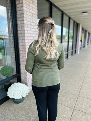 Woman wearing olive green ribbed knit long-sleeve top with dark pants, walking down covered walkway
