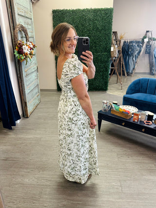 Woman wearing a floral midi dress with off-shoulder puff sleeves, smiling in boutique mirror selfie.