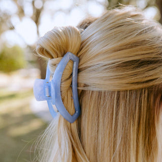 Blonde woman wearing a blue open hair clip holding her hair in a ponytail outdoors.