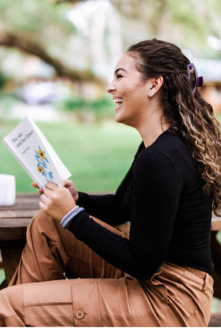 Woman with curly hair wearing burgundy hair clip, smiling while reading a book outdoors.