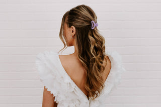 Woman wearing lilac hair clip in long wavy blonde hair, shown from behind in white top.