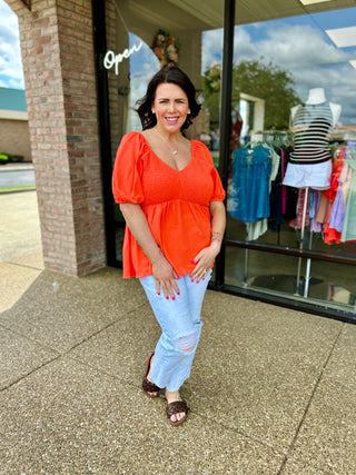 Woman wearing orange smocked peplum top with puffy sleeves standing outside clothing boutique storefront