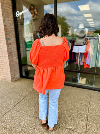 Woman wearing orange smocked peplum top with puffy sleeves and light blue jeans, shown from behind.