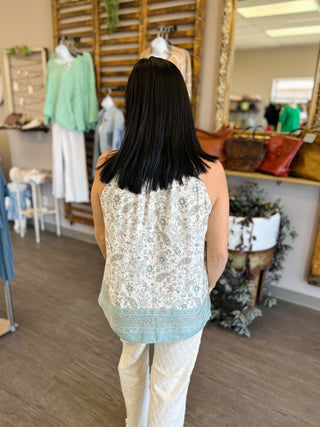 Woman wearing paisley print sleeveless top with decorative border, viewed from behind in boutique setting.