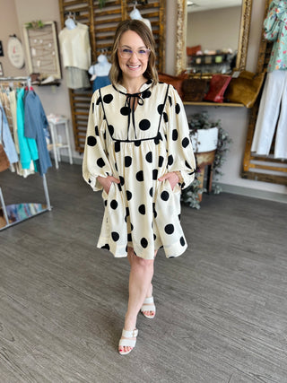 Woman wearing cream polka dot dress with black trim and pockets in clothing boutique setting.