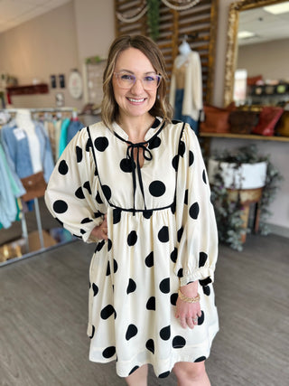 Woman wearing cream polka dot dress with black tie neck and long sleeves in boutique setting