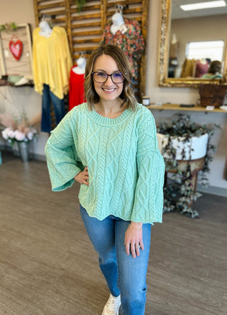 Woman wearing mint green cable knit sweater with jeans in boutique clothing store setting