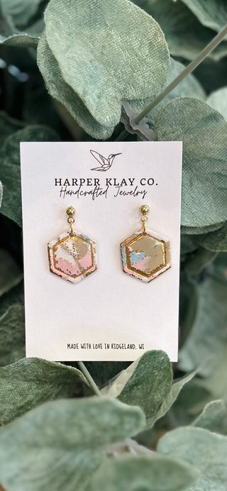 Hexagonal dangle earrings with pastel marble pattern and gold trim displayed on jewelry card among green leaves.
