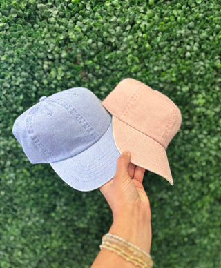 Hand holding two pastel-toned unstructured baseball caps against a green leaf wall background.