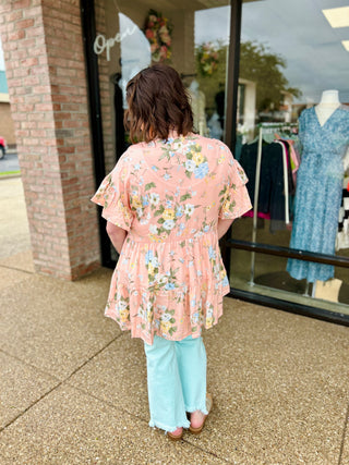 Woman wearing floral kimono and jeans standing outside boutique storefront, viewed from behind.