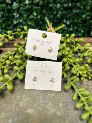 Two pairs of pear-cut stud earrings on white cards surrounded by green foliage on wooden surface.